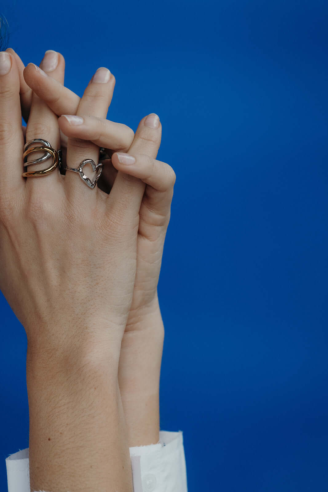 Close-up of clasped hands against a blue background, one wearing the open silver heart-shaped Street Side RING stacked with gold rings.