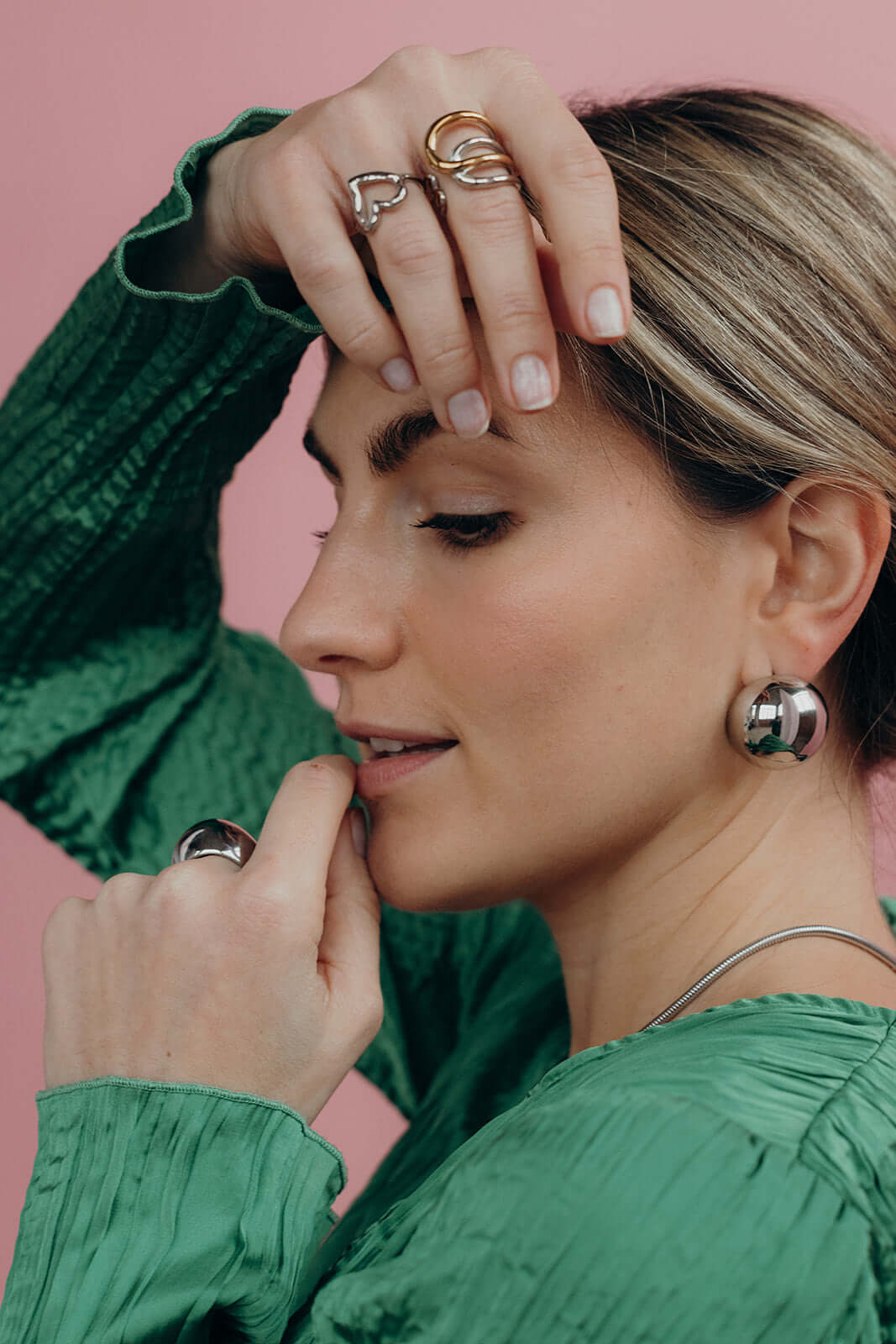 Close-up of a hand near the face wearing the silver heart-shaped Street Side RING stacked with other rings, pink backdrop.