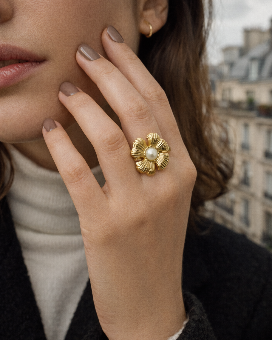 Close-up of a hand near the face wearing the gold textured flower Summertime Harmony RING with its pearl center, city backdrop.