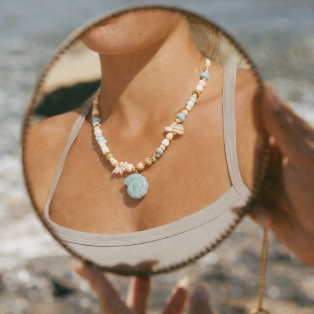 The Tahiti NECKLACE worn at the beach, reflected in a round hand mirror as the model shows off its carved amazonite shell pendant.