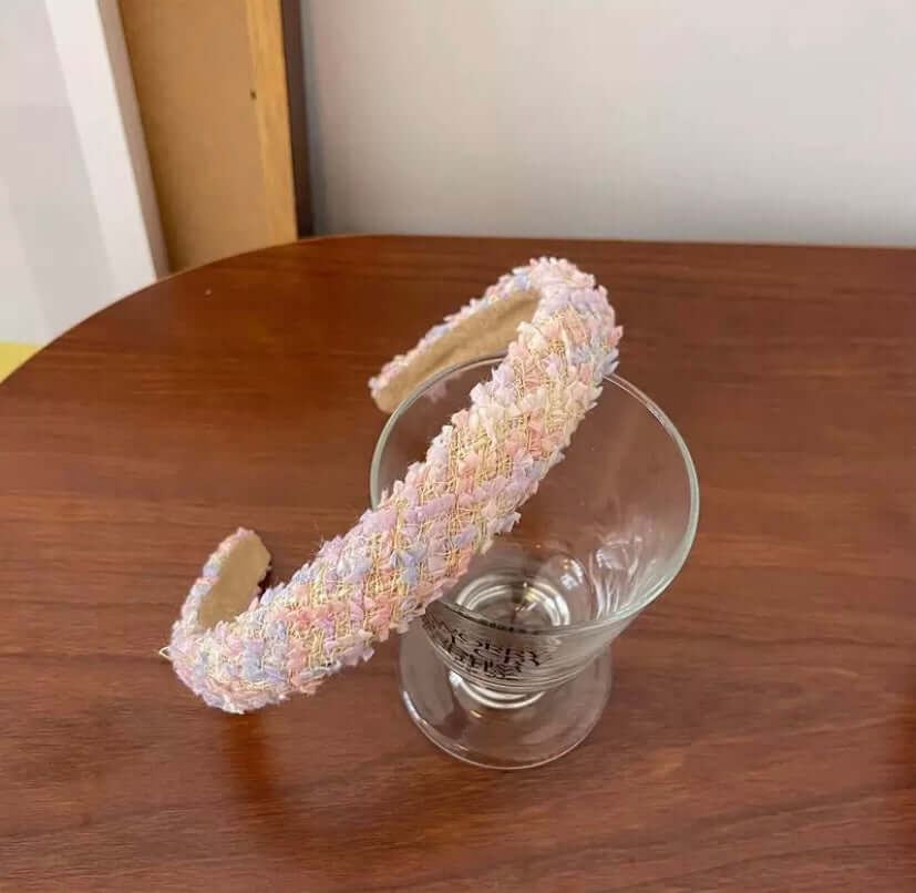 A single pastel multicolor tweed headband in pink, blue and yellow resting on a wood table beside a glass.