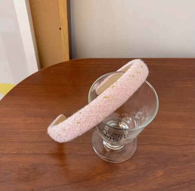 A single blush pink boucle headband with gold fleck detail resting on a wood table beside a glass.
