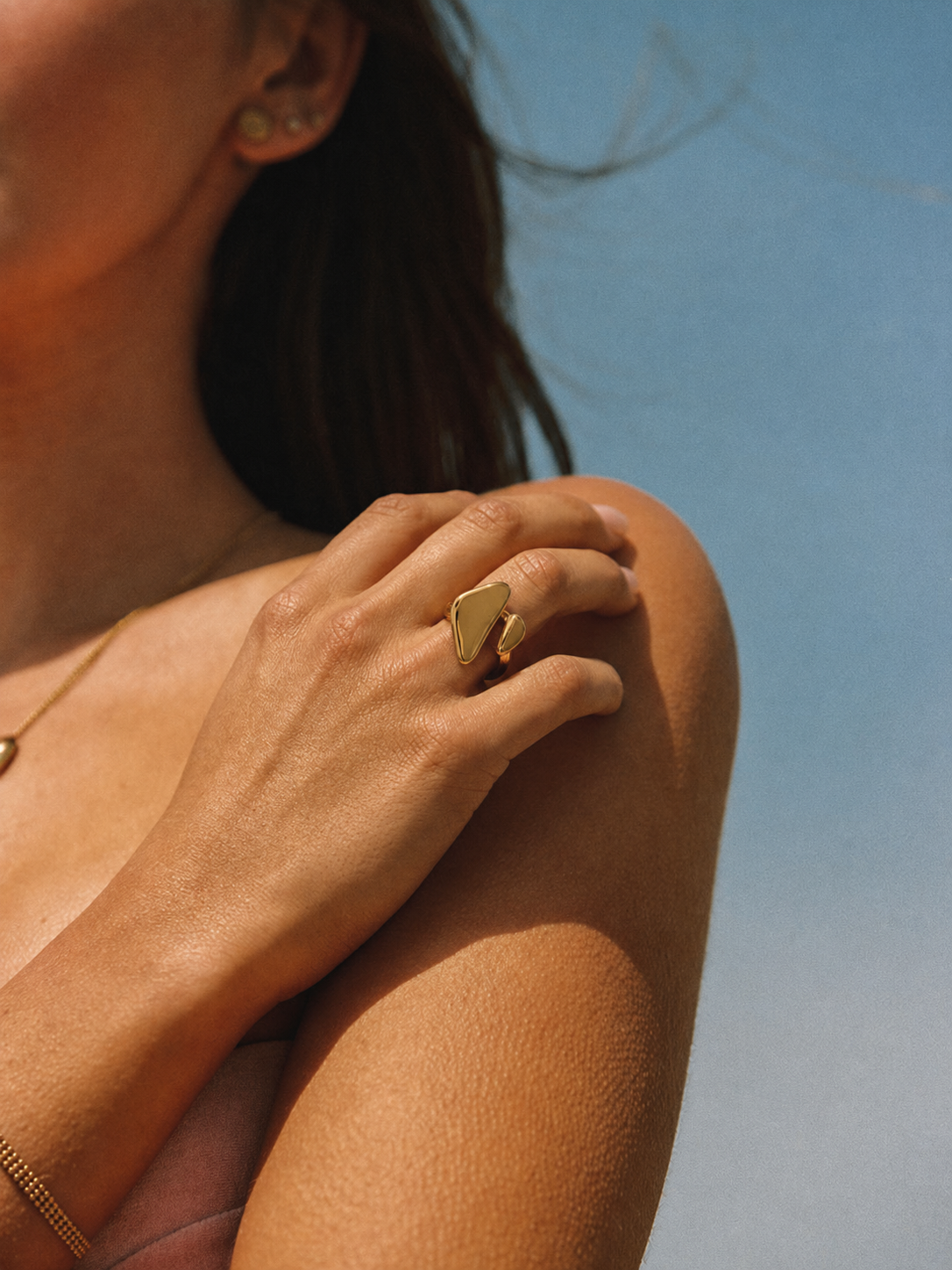 Model wears the gold Vénus RING's triangular face on her shoulder, hair blowing against a clear blue sky.