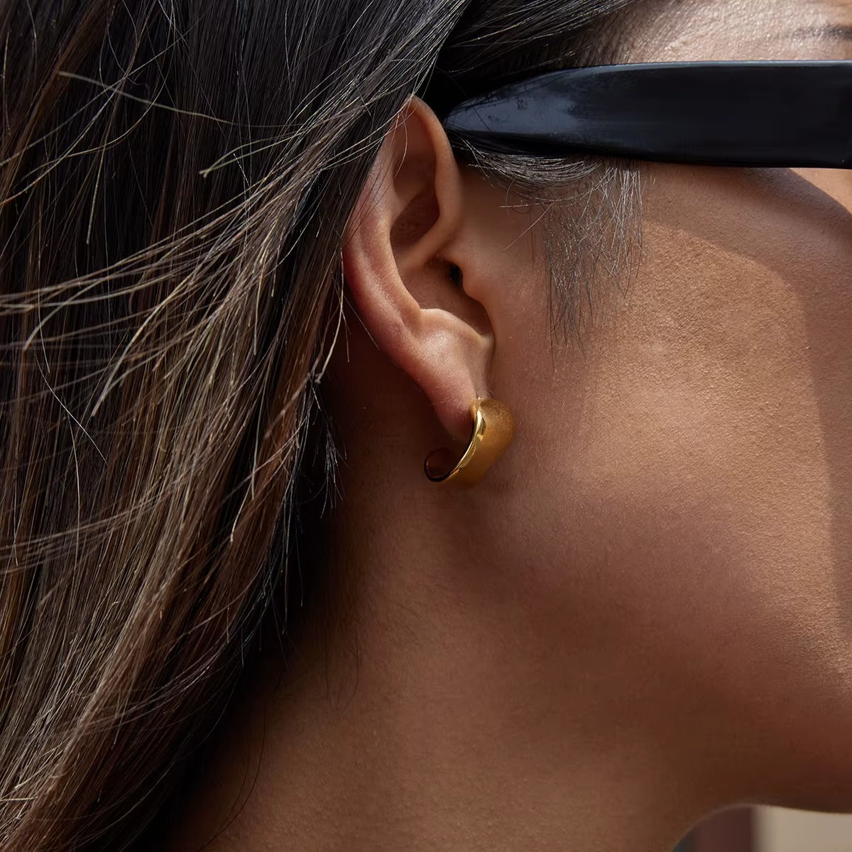Verbier Earring worn on the ear in gold, styled with sunglasses, the wave-shaped hoop shown from behind.