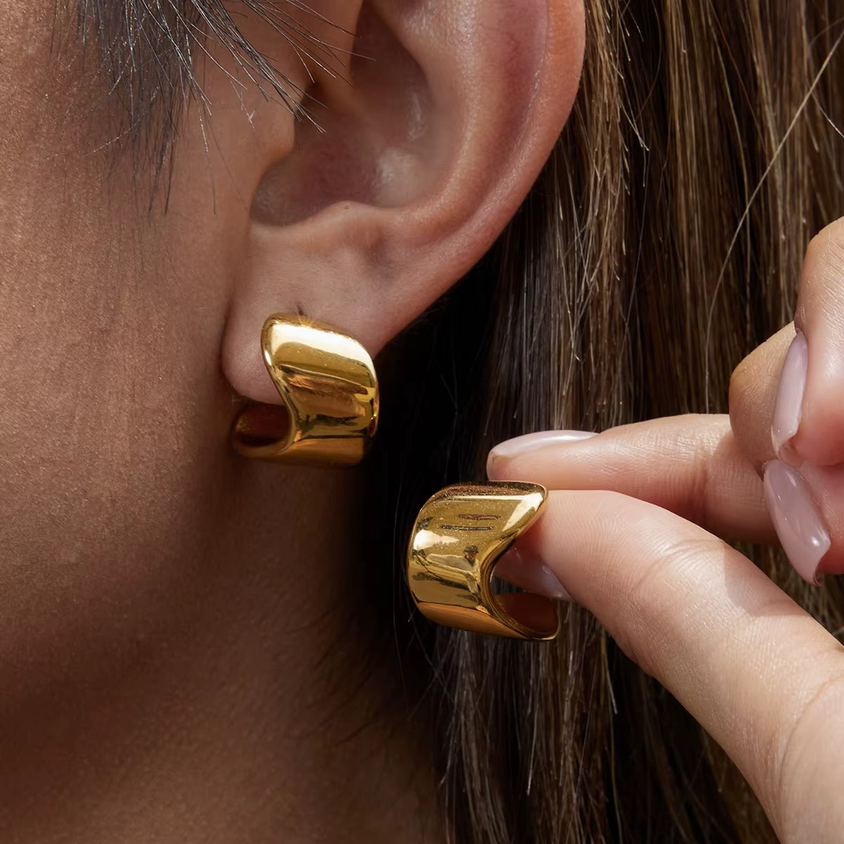 Verbier Earring worn on the ear in gold, a hand reaching up to gently touch the sculptural wave hoop.