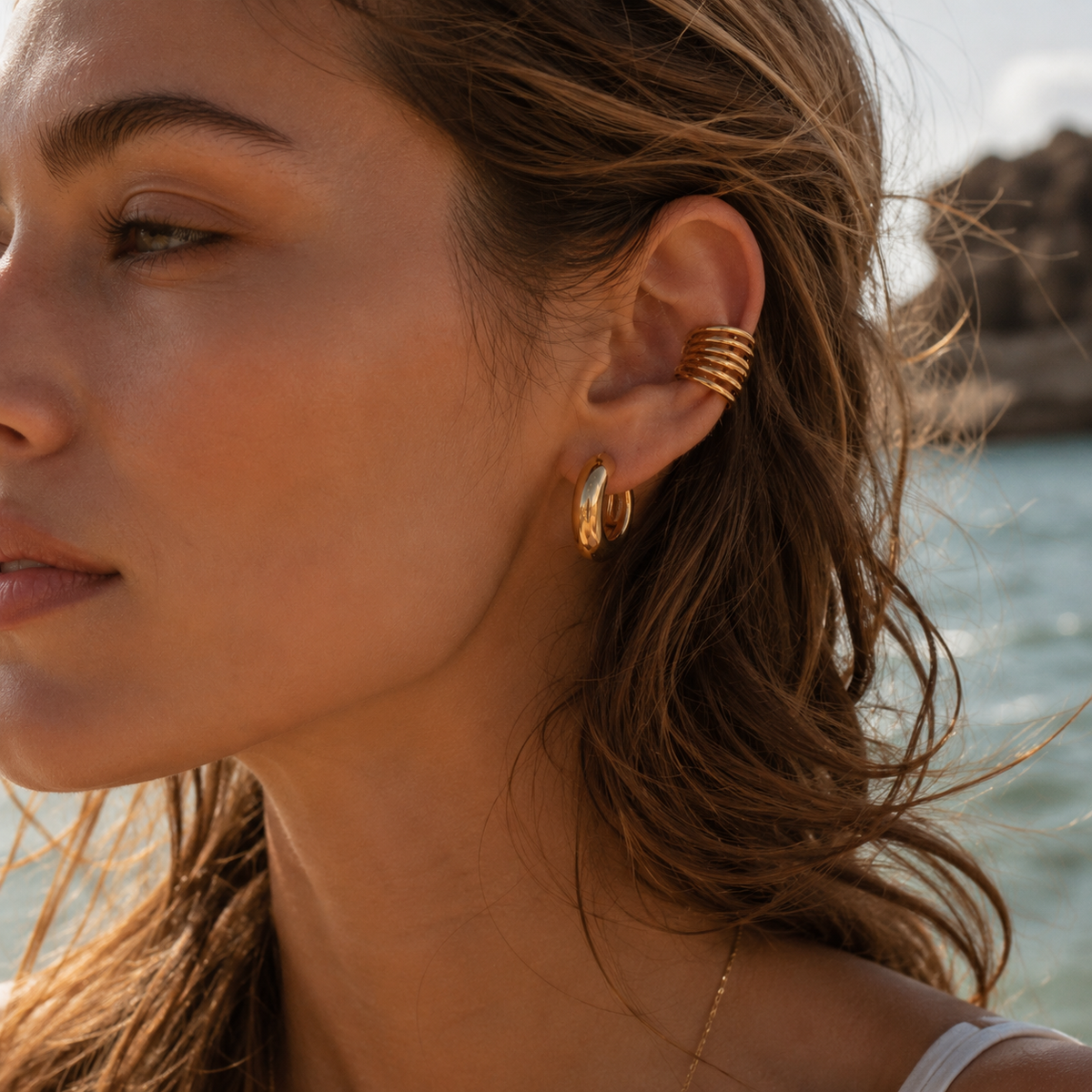 The Whitney EAR CUFF worn on the ear at the beach, a multi-band gold cuff layered above matching gold hoop earrings.