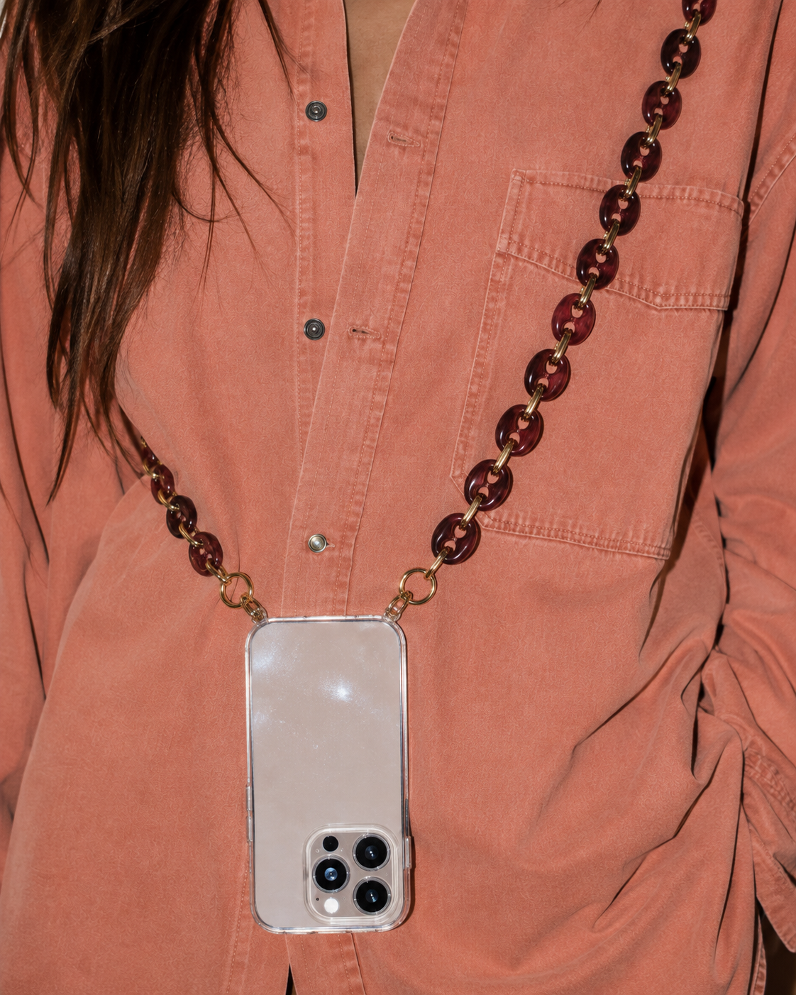 Model wearing a rust-colored shirt with a phone in a clear case suspended from the Folie à Trois Wilder phone chain in wine red puffed links.
