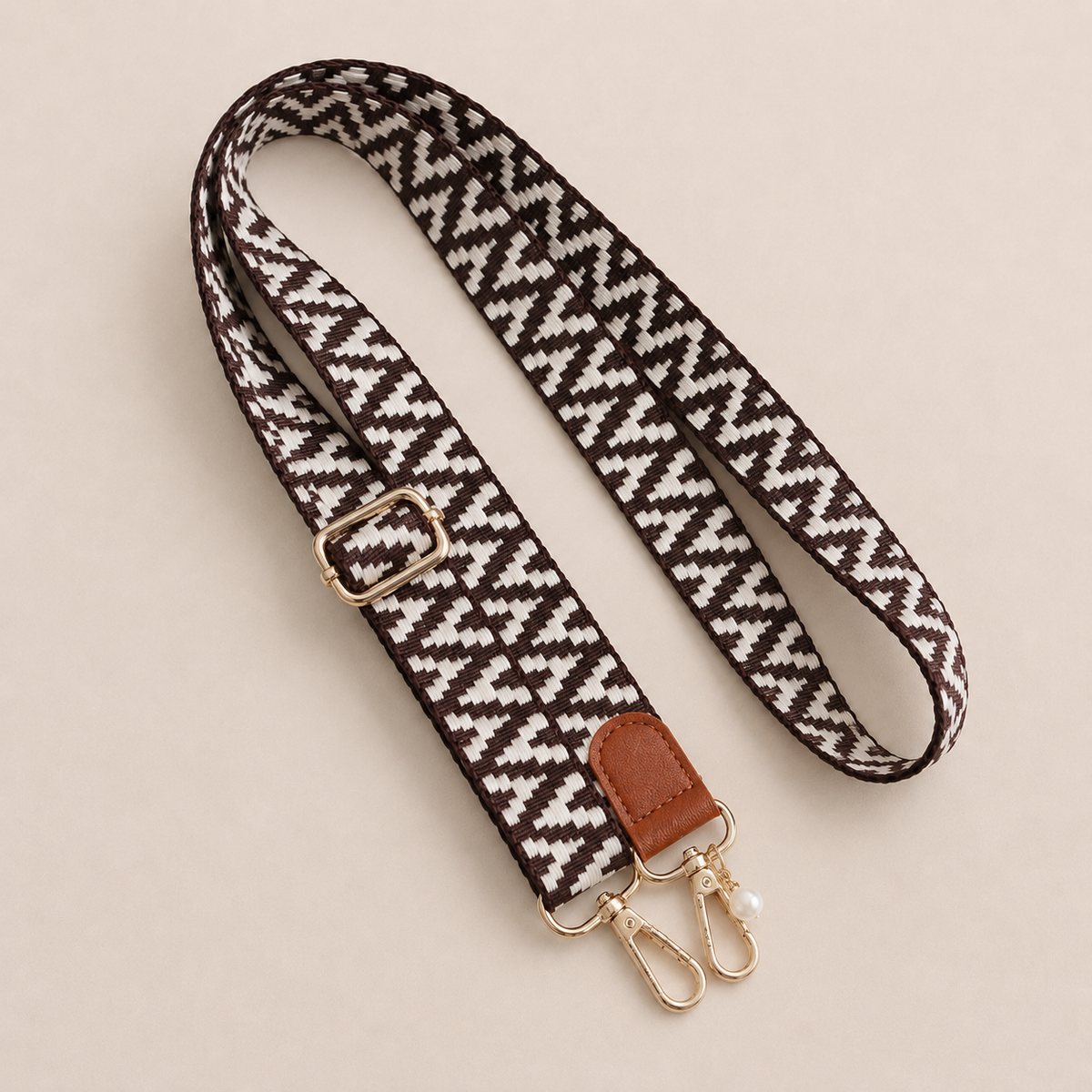 Folie à Trois Wynter long wholesale phone strap in a brown and white zigzag weave with a tan leather tab, gold clasps and a pearl charm, coiled on a cream background.