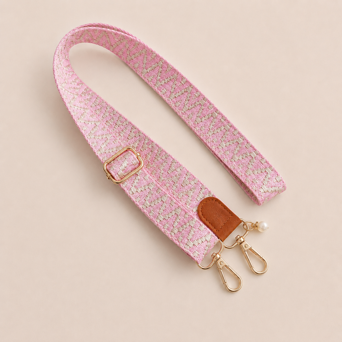 Folie à Trois Wynter long wholesale phone strap in a pink and beige zigzag weave with a tan leather tab, gold clasps and a pearl charm, coiled on a cream background.