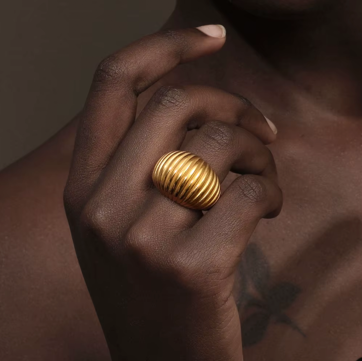 Close-up of a hand near the collarbone wearing the chunky gold ribbed Zelma RING, warm dramatic lighting.