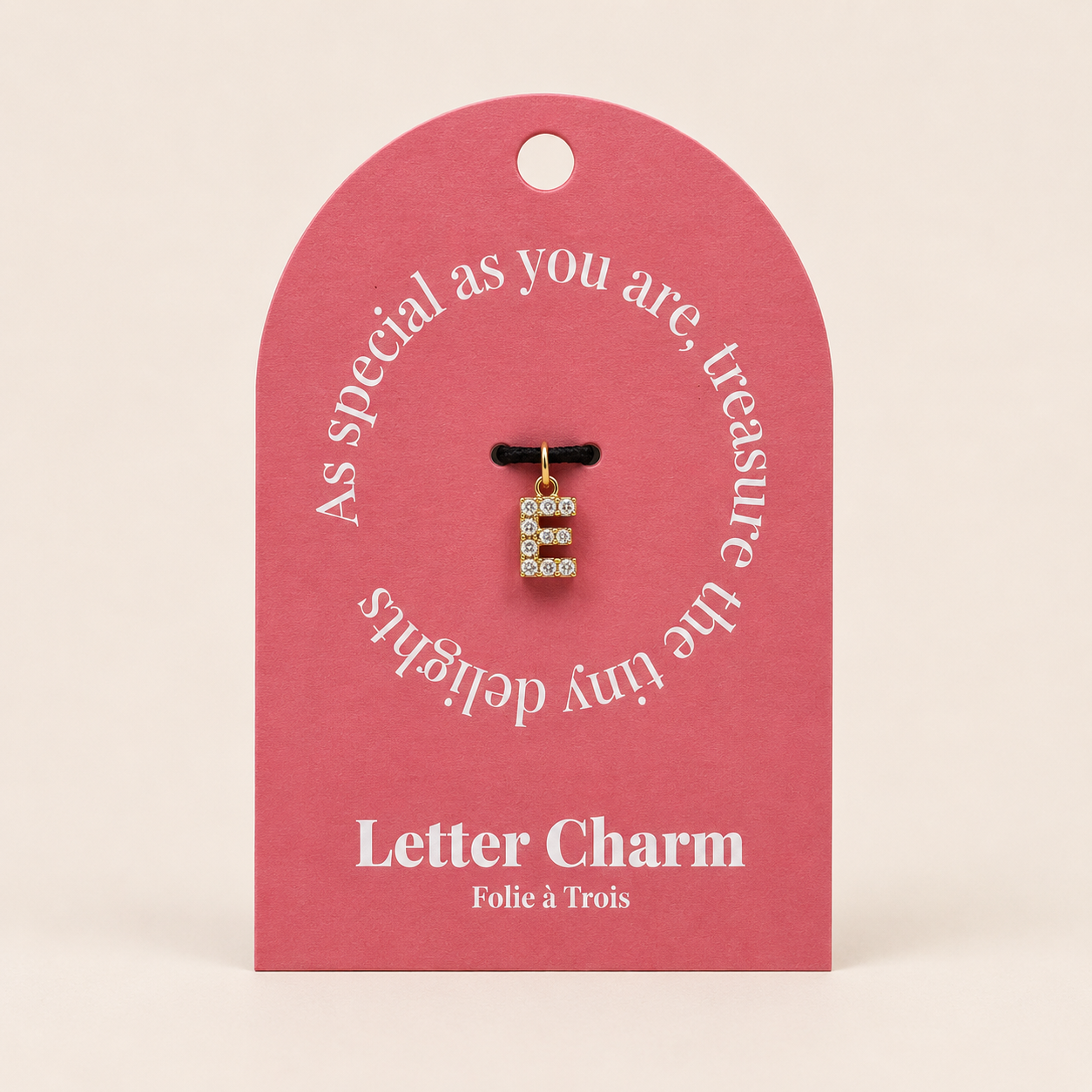The Zircon Letter CHARM in E, a zircon-set gold initial displayed on a pink Letter Charm gift tag with a black cord.