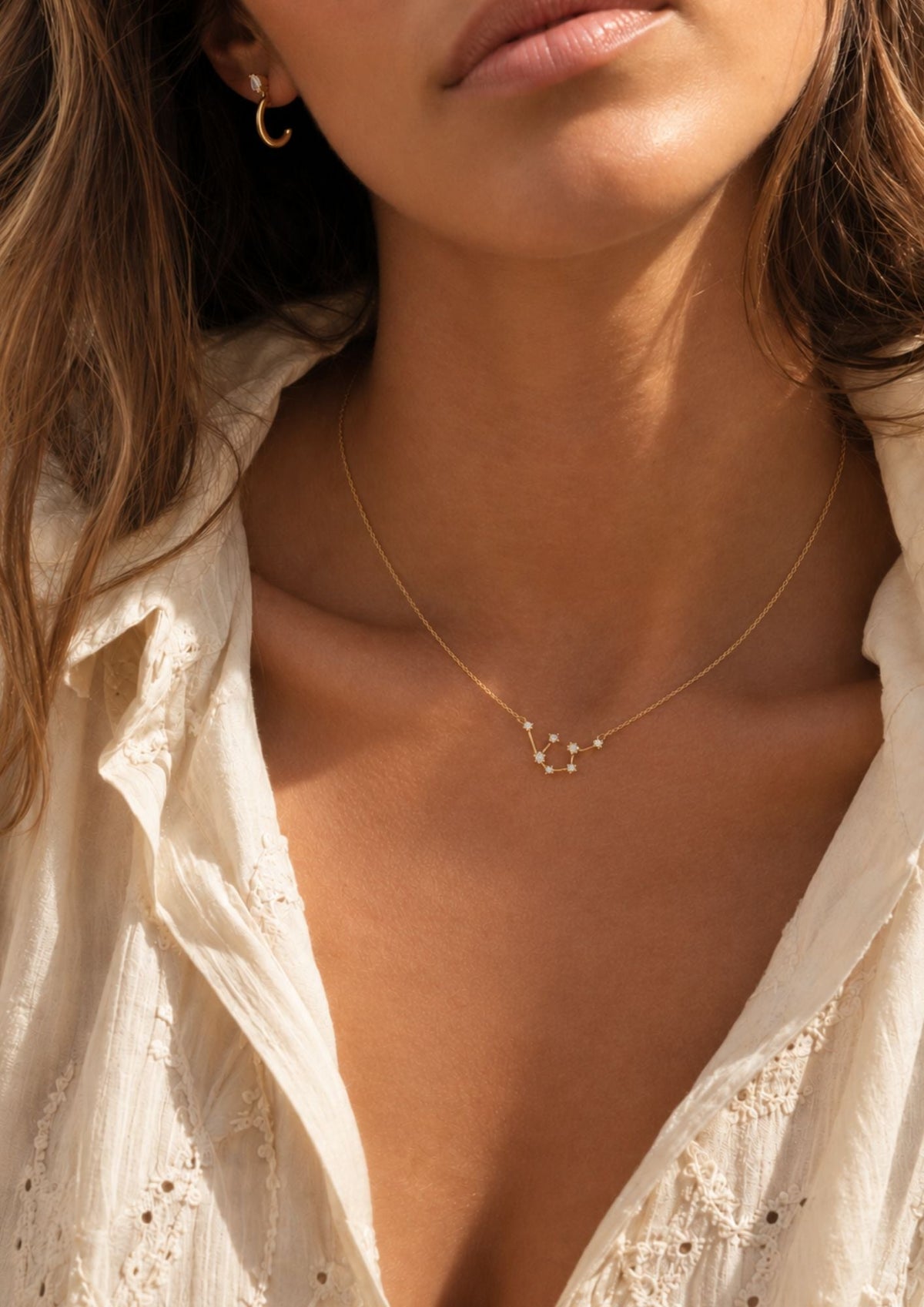 Sunlit close-up of a model in a white eyelet blouse wearing the gold Zodiac NECKLACE with a crystal constellation pendant.
