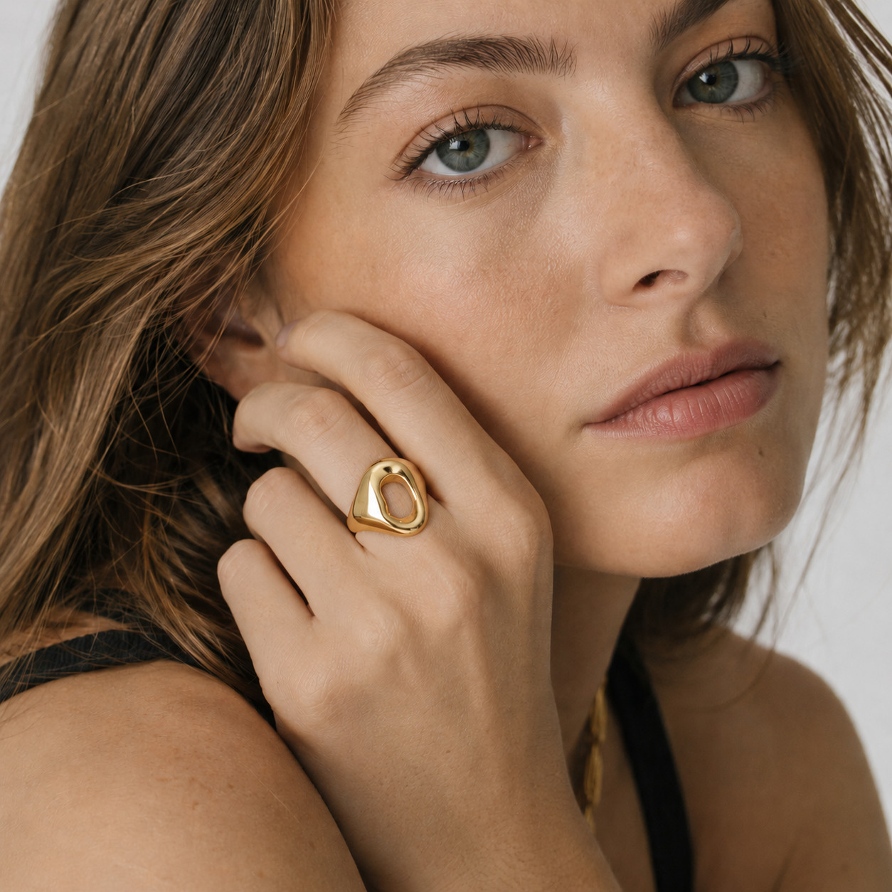 A close-up lifestyle shot of a woman wearing a chunky, polished gold ring on her finger, with her hand resting against her cheek.