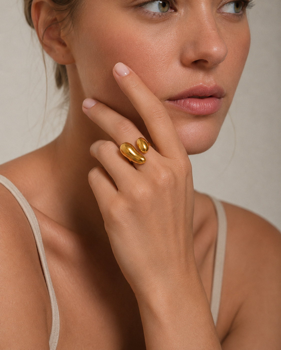A close-up lifestyle shot of a model wearing a chunky, sculptural gold ring on her finger, with her hand resting against her cheek.