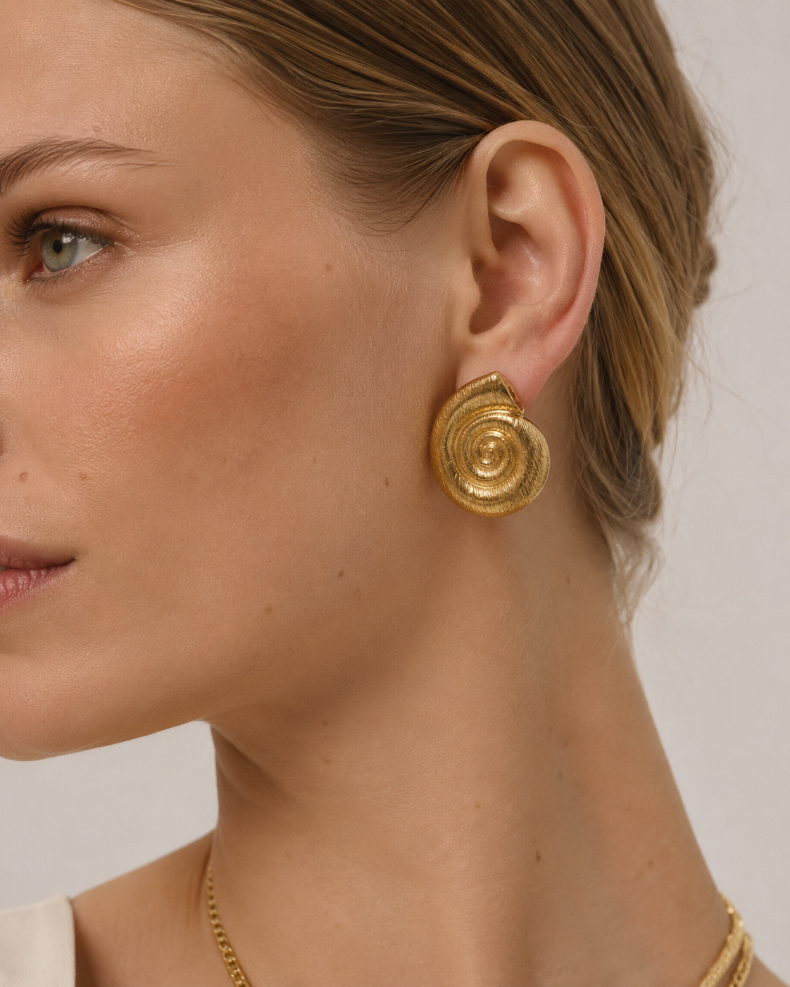 A close-up of a woman's ear wearing a textured gold spiral drop earring, with her hair pulled back and a delicate gold chain necklace visible against a light neutral background.