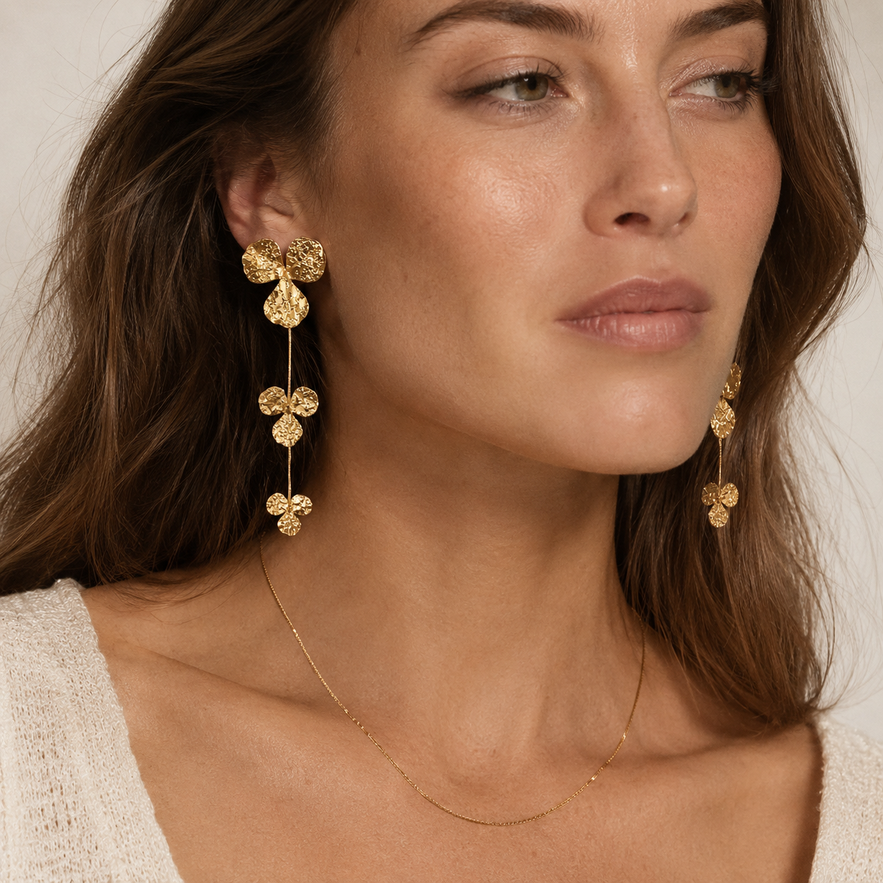 A close-up of a woman wearing a pair of gold dangle earrings with textured circular drops and a delicate gold necklace, set against a soft beige background.