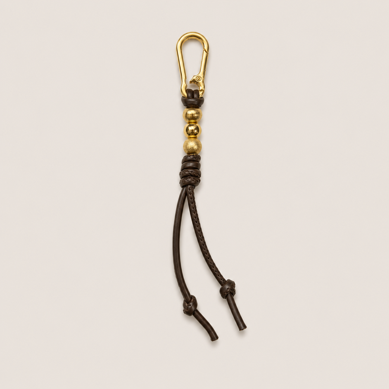 Taupe braided bag charm with a gold-tone carabiner clip, decorative beads and knotted tassels on a light beige background.