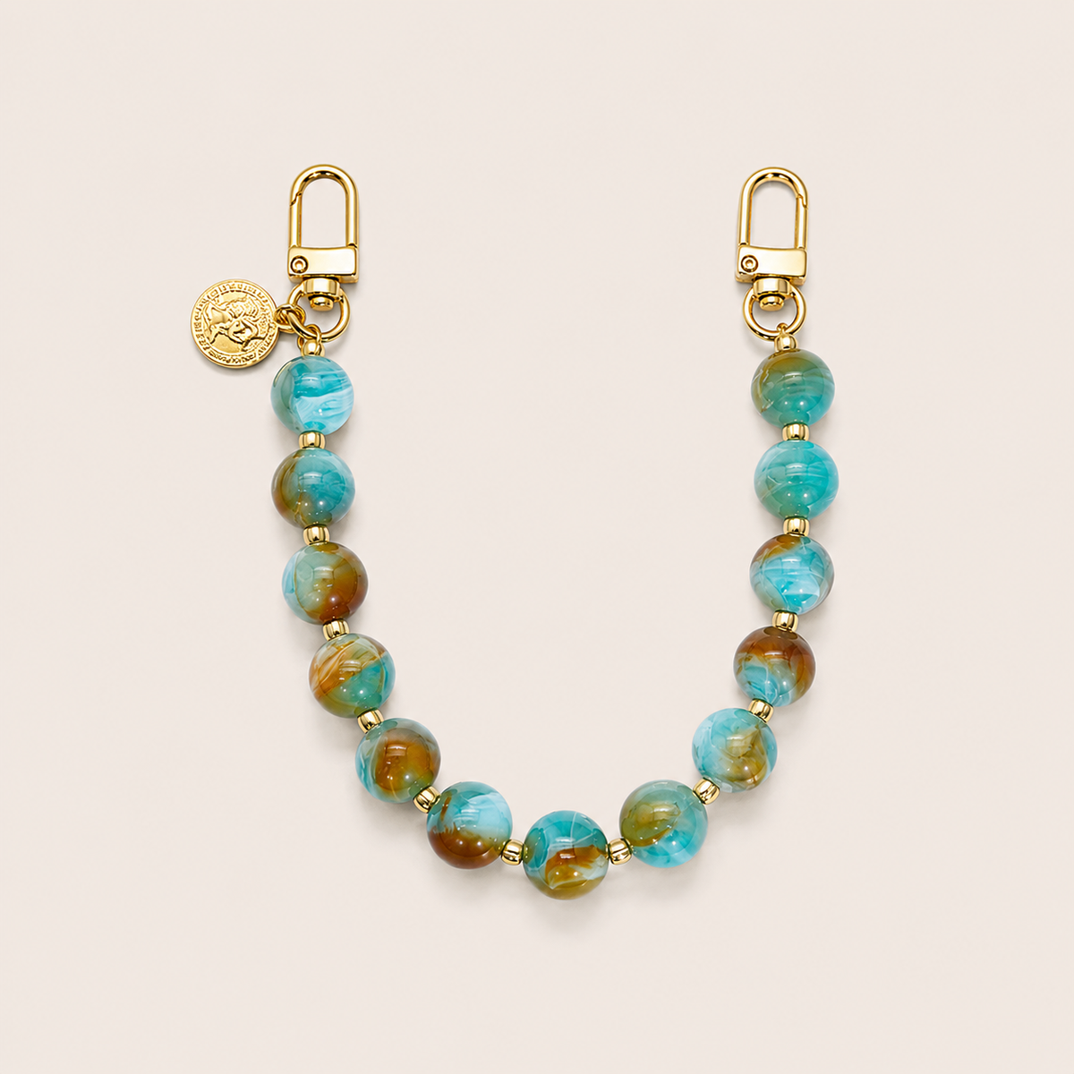 The Sable PHONE CHAIN in turquoise and brown marbled beads with gold-tone clasps and a small round gold coin charm, on a light beige background.