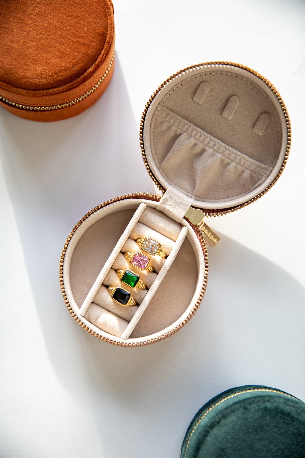 Open round Travel Jewelry CASE in blush velvet with a ring roll holding gold gemstone rings, cognac and green cases nearby.
