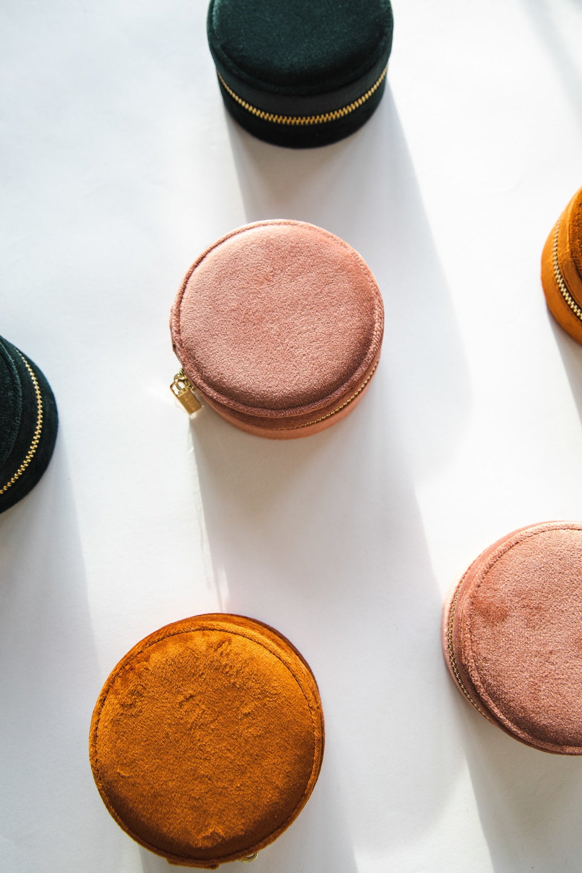 Round Travel Jewelry CASE pouches in blush pink, cognac and dark green velvet scattered in sunlight on a white surface.
