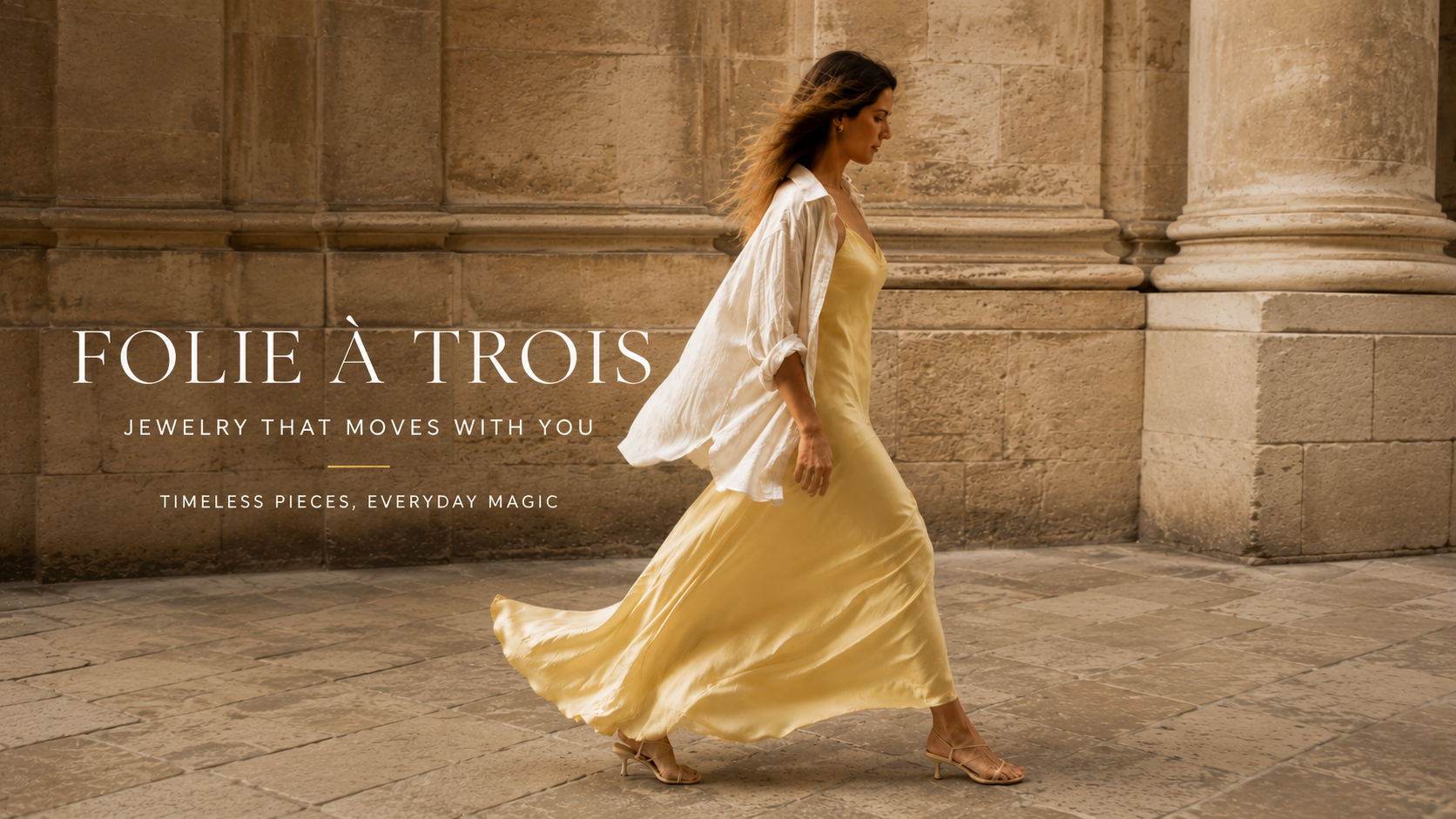 Woman in a flowing yellow dress walking through a stone archway, with text reading "Folie à Trois, jewelry that moves with you."