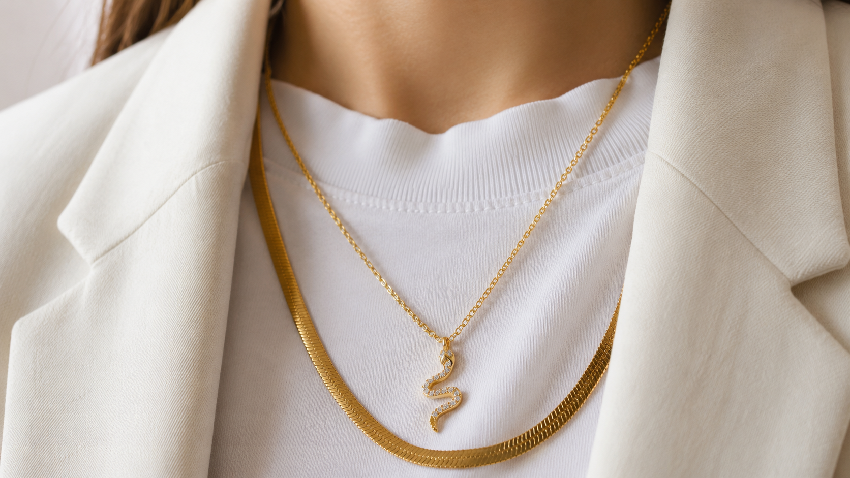 Close-up of a woman's collarbone in a white top and cream blazer, wearing delicate layered gold necklaces.