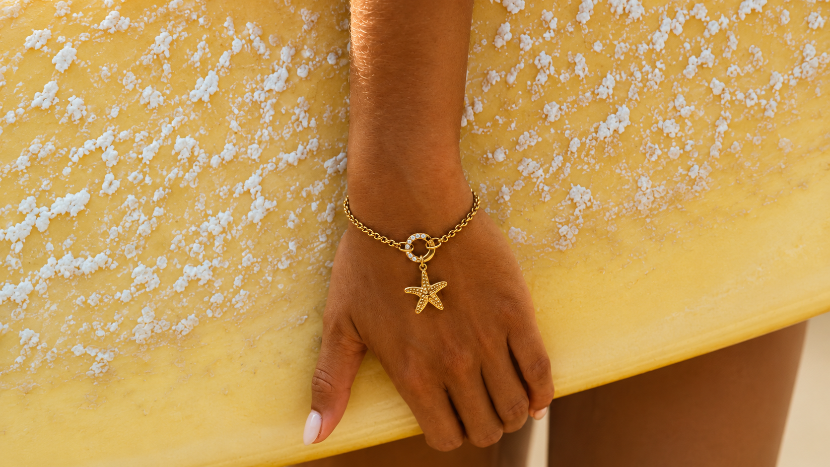 Close-up of a tanned hand resting on a yellow towel, wearing a gold chain bracelet with a starfish charm.