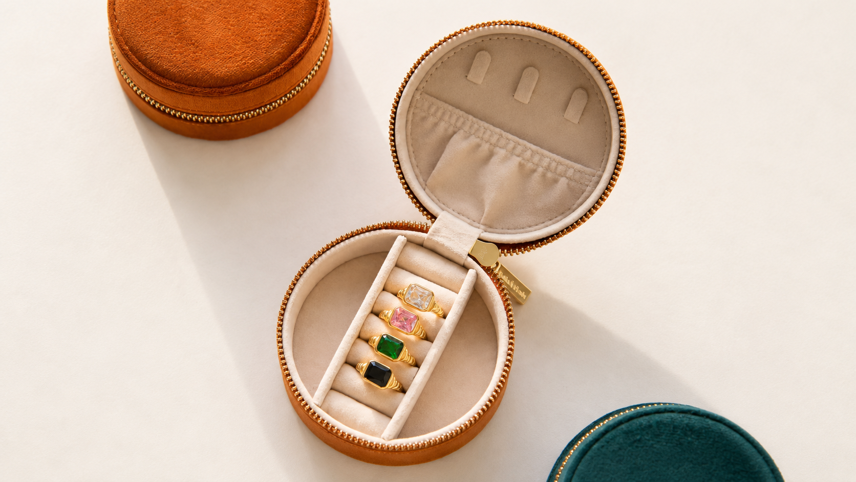 Flat lay of round leather jewelry travel cases in orange, cream and teal, one open showing rings inside.