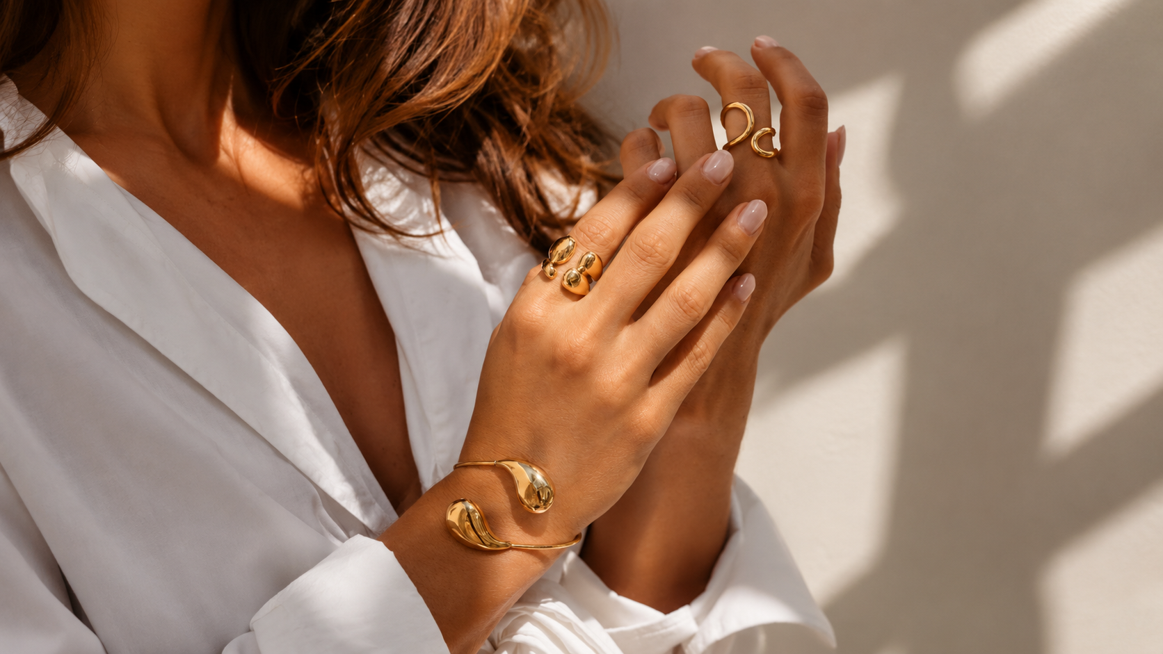 Close-up of two hands in sunlight wearing white shirt sleeves, stacked with layered rings and gold bangle bracelets.