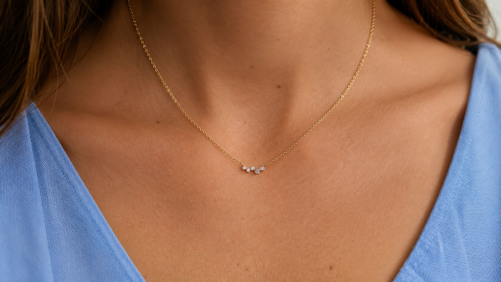 Close-up of a woman's neckline in a blue top, wearing a delicate fine chain necklace with a small pendant.