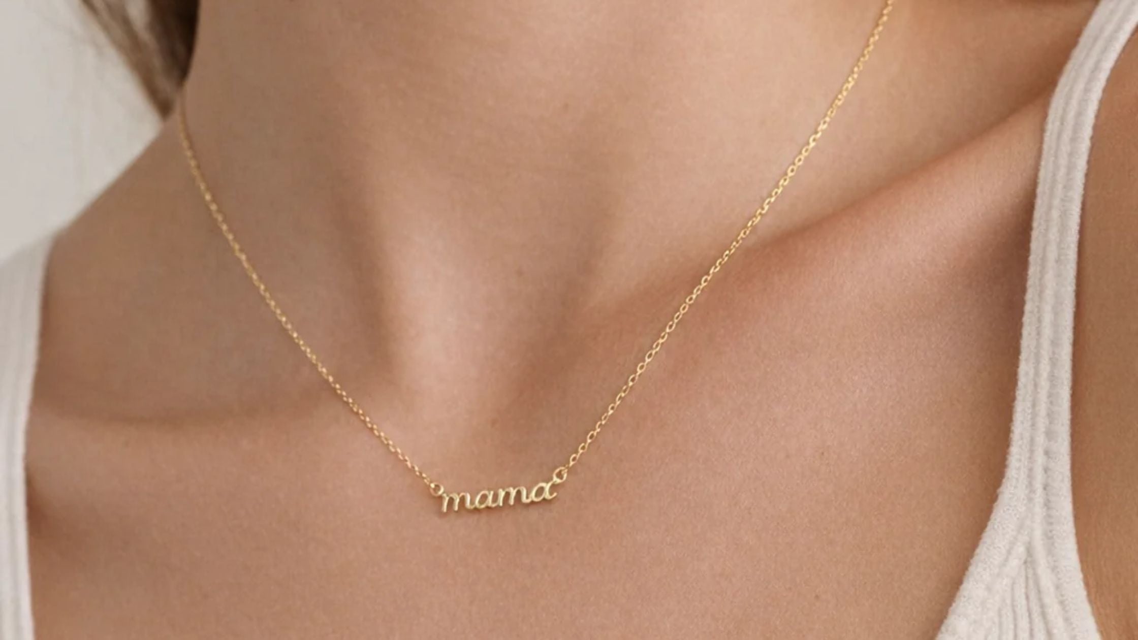 Close-up of a woman's neckline in a white top, wearing a gold necklace with a script name pendant.