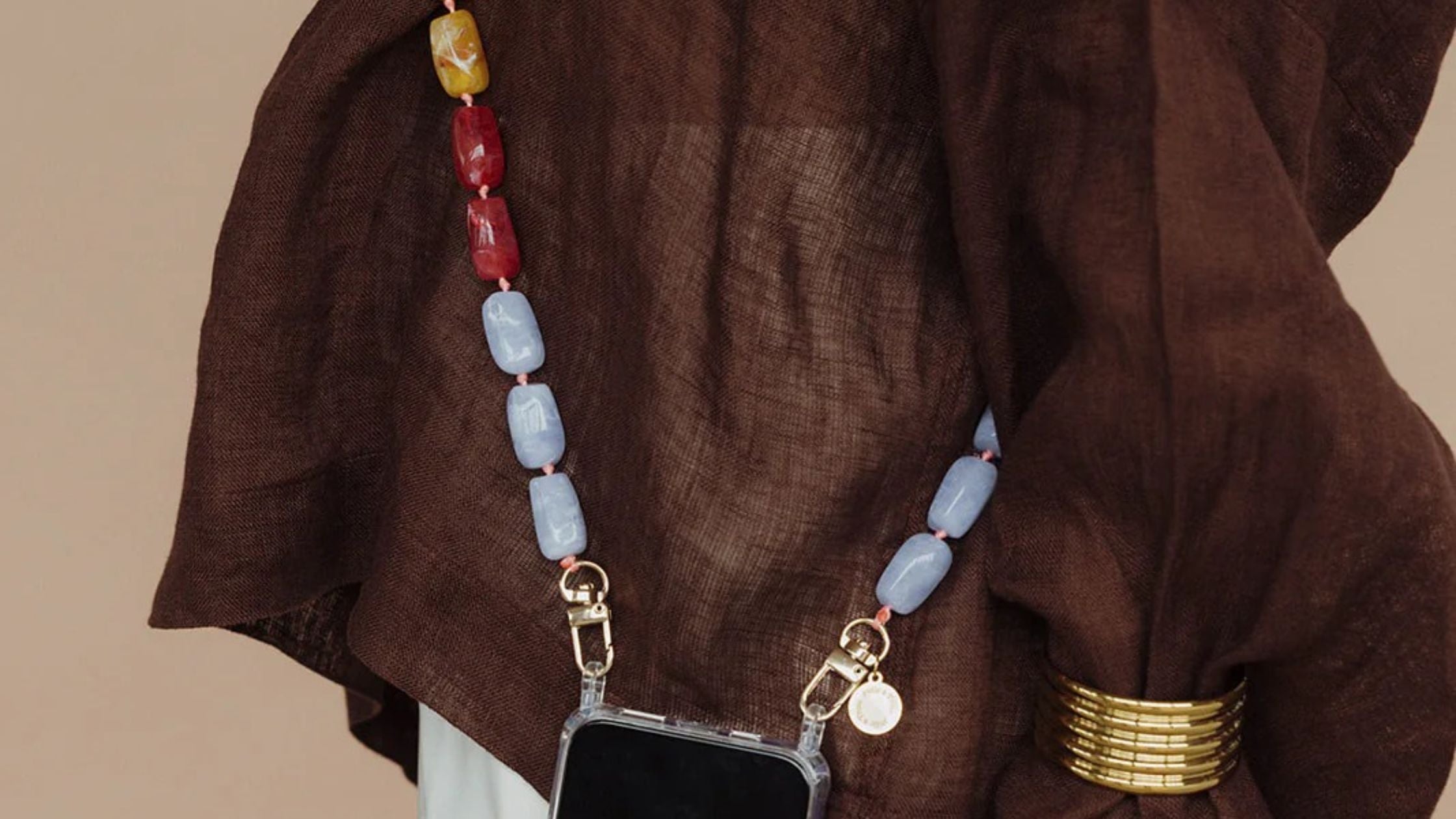 Close-up of a woman's wrist in a brown outfit wearing a gold bracelet, holding a phone with a beaded charm strap.