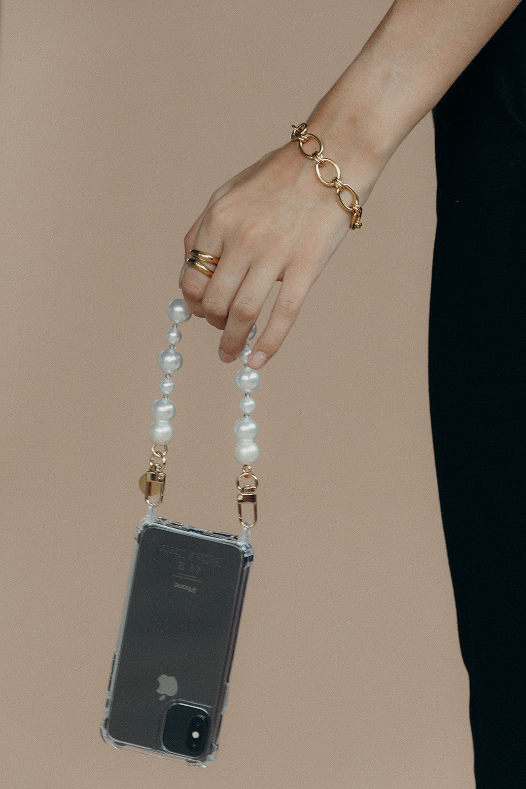 Model's hand holding a phone in a clear case by the Folie à Trois Adair Pearl wholesale phone chain against a black outfit.