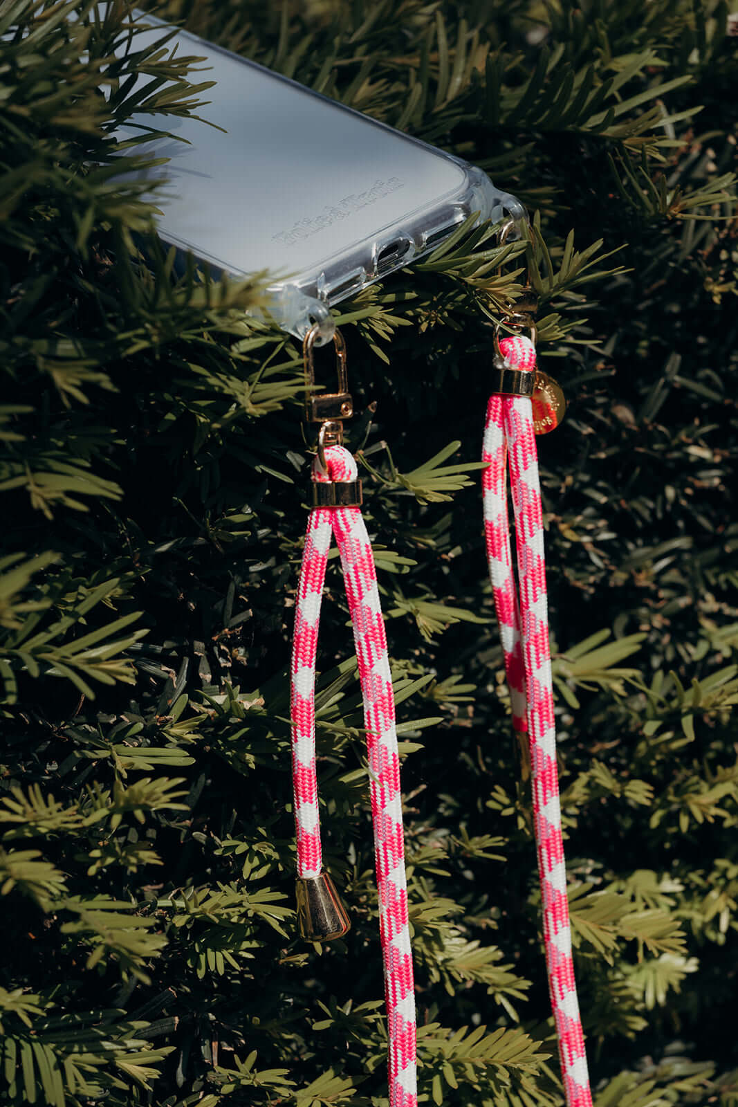 Close-up of a clear phone case with pink and white Aesthetic Colorful PHONE CORD clasps hanging in greenery.