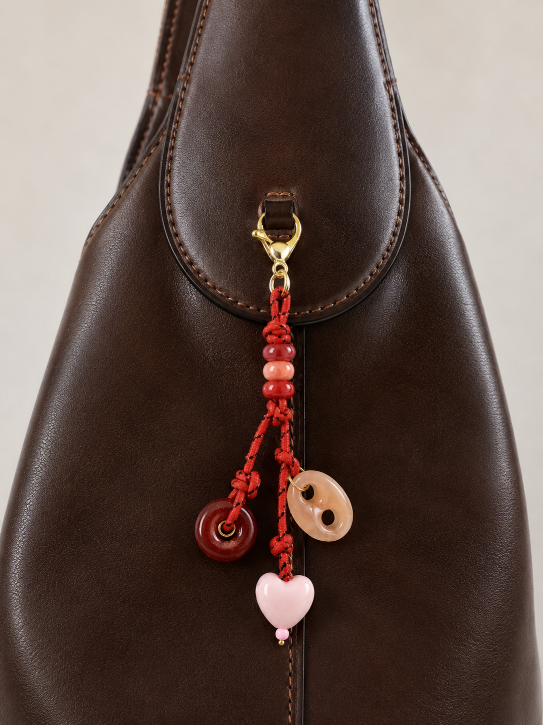 Folie à Trois Amara BAG CHARM, a red beaded cord with heart and agate donut charms, clipped to a dark brown leather bag.