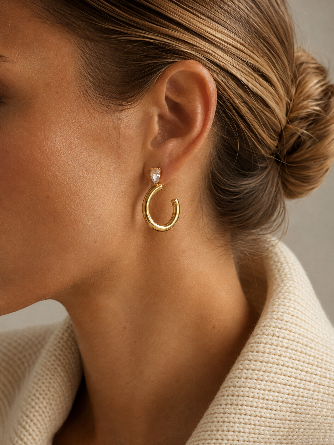 Close-up of a model in a cream coat wearing the gold Aurora EARRINGS, a chunky tube hoop topped with a marquise zirconia.