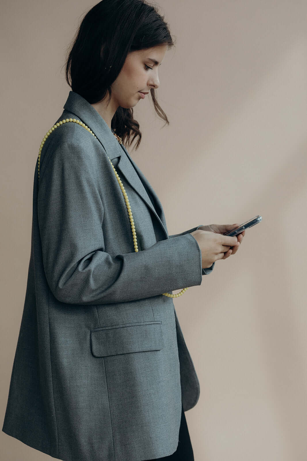 Woman in a gray blazer using her phone with a pale yellow beaded Aurora PHONE CORD draped over her shoulder.