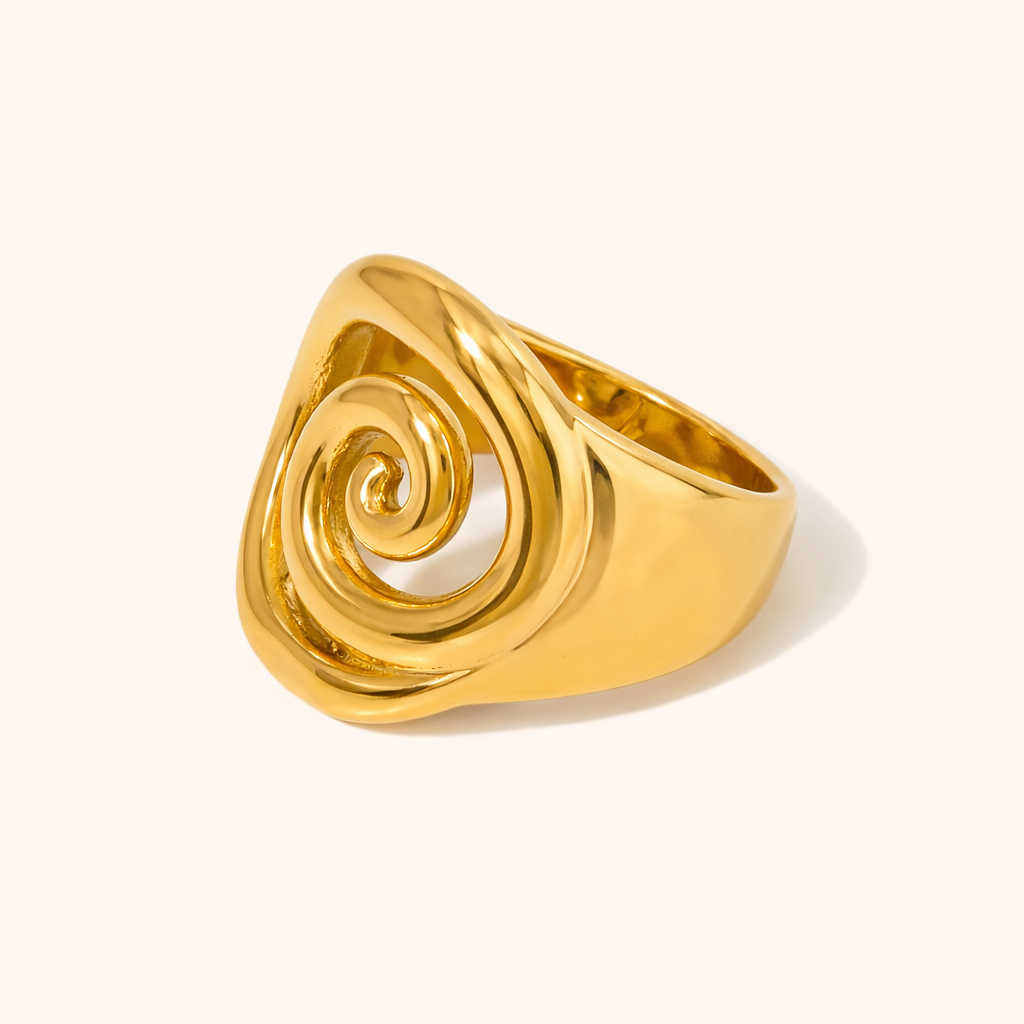 Gold signet-style ring with a spiral swirl carved into the wide oval face, shown at an angle on a cream backdrop — Avery RING.
