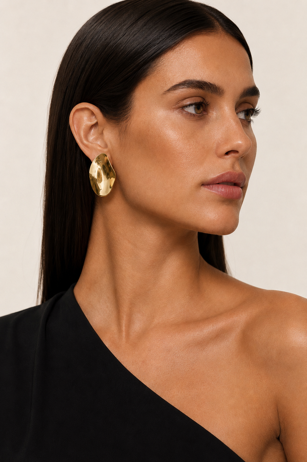 Model in a black one-shoulder top wears the gold Axis EARRINGS, a sculptural hammered dome catching the light at the ear.