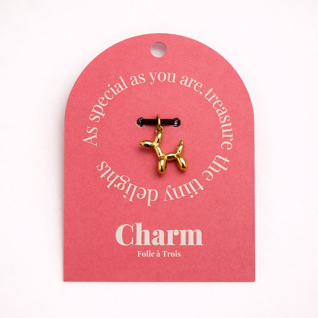 The Balloon Dog CHARM displayed on a coral pink Charm gift tag, the gold balloon-dog pendant hanging from a black cord.