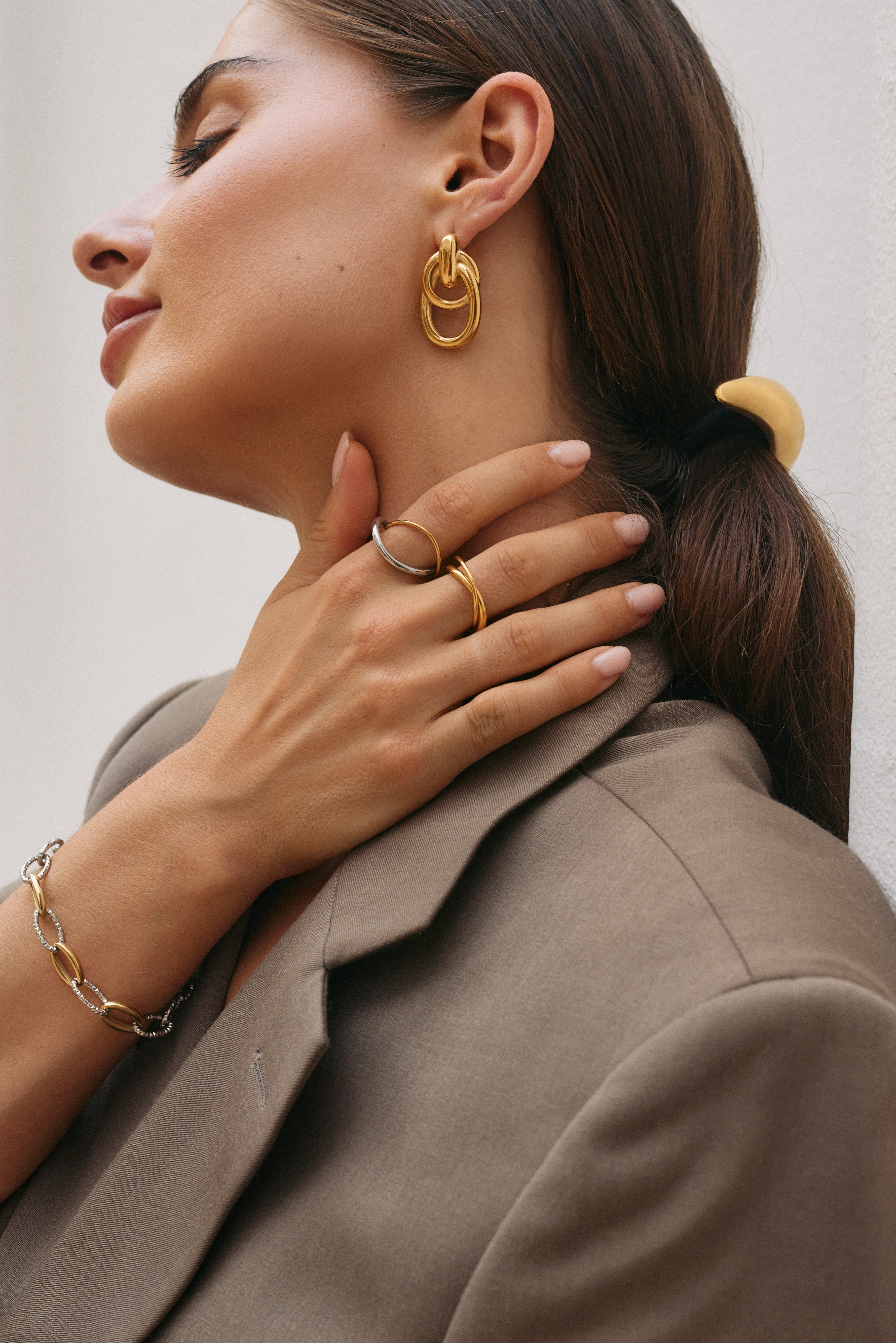 Close-up of a hand near the neck stacked with the silver interwoven Belance RING and a gold band, matching hoop earring visible.