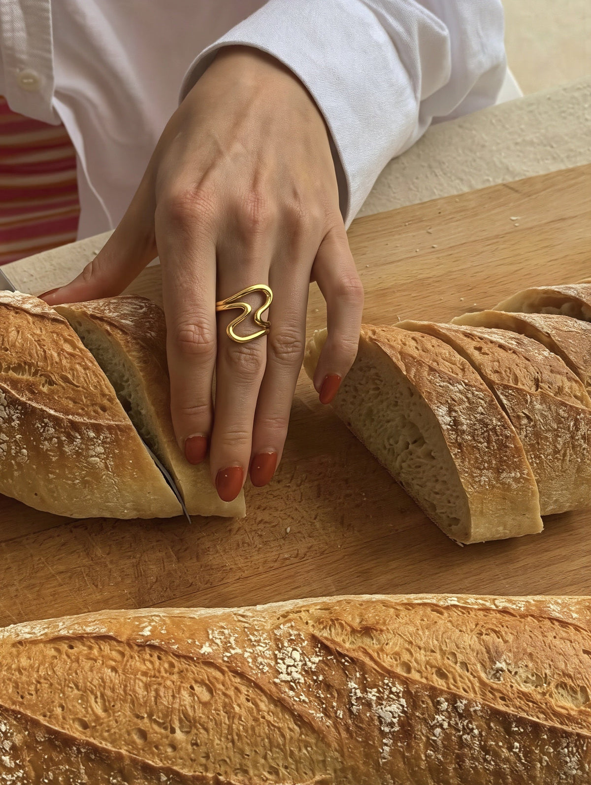 Hand wearing the gold wave-shaped Big Wave RING while slicing fresh bread on a wooden cutting board, a warm everyday lifestyle shot.
