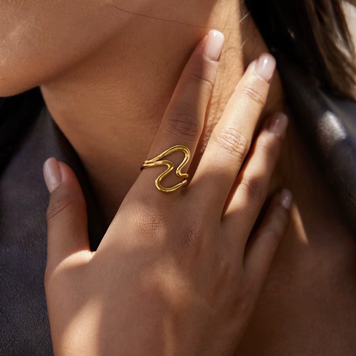 Close-up of a hand raised near the collarbone wearing the gold Big Wave RING, dark leather jacket visible in the background.