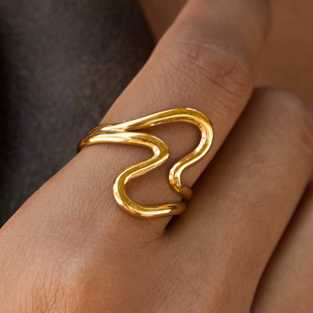 Extreme close-up detail shot of the gold Big Wave RING on the hand, showing its wavy interlocking loop design up close.