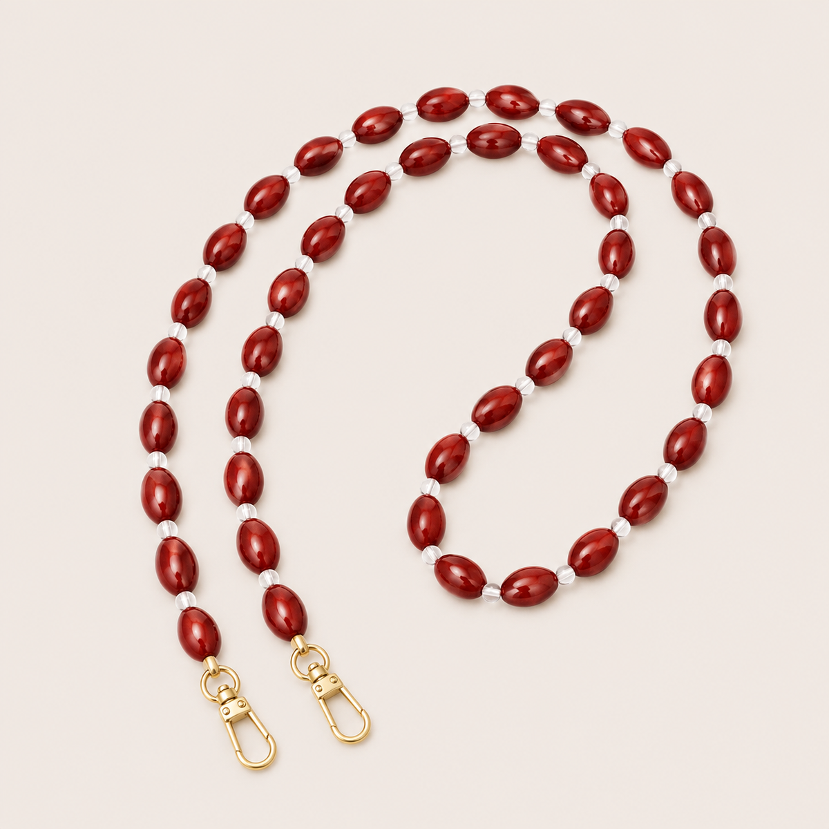 Folie à Trois Bramble long phone chain in deep red oval beads with clear spacers and gold clasps, coiled on a cream background.