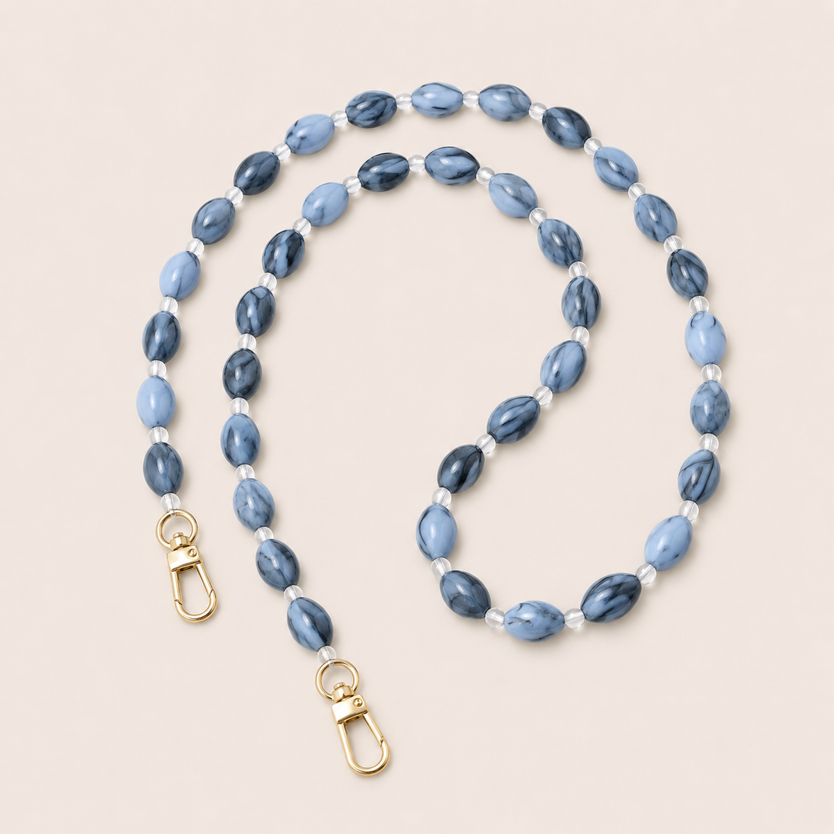Folie à Trois Bramble long phone chain in blue marbled oval beads with clear spacers and gold clasps, coiled on a cream background.
