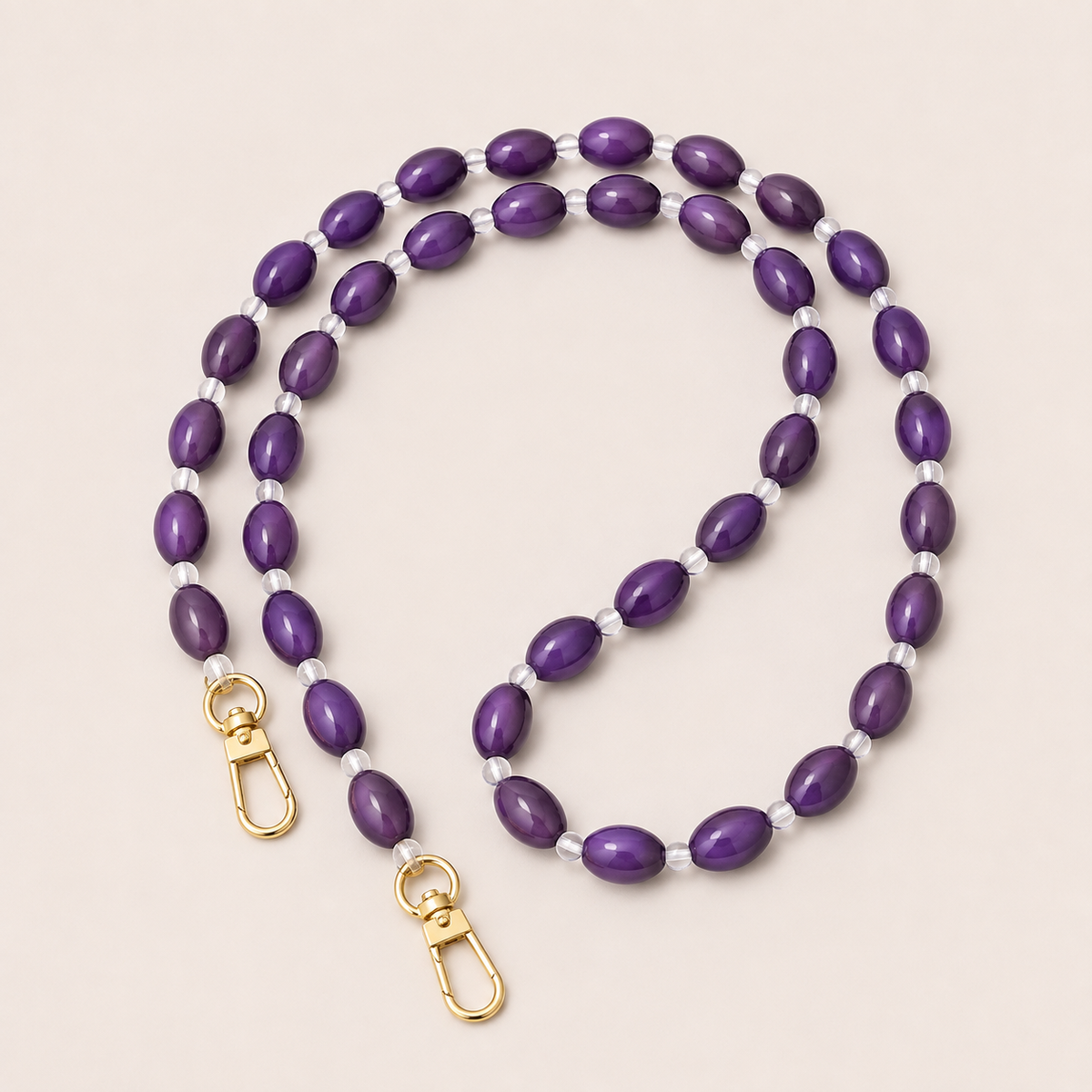 Folie à Trois Bramble long phone chain in purple oval beads with clear spacers and gold clasps, coiled on a cream background.