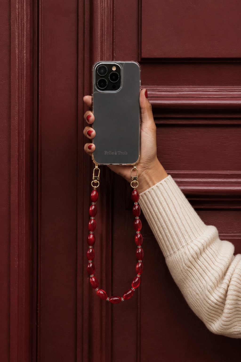 Model holding a clear phone case attached to the Folie à Trois Bramble wholesale phone chain in deep red beads, against a red paneled door.