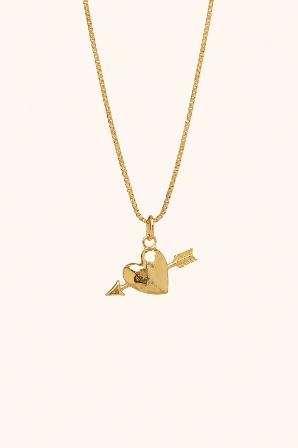 The Burning Hearts NECKLACE in gold, a puffed heart pierced by an arrow charm, hung from a box chain on a cream background.
