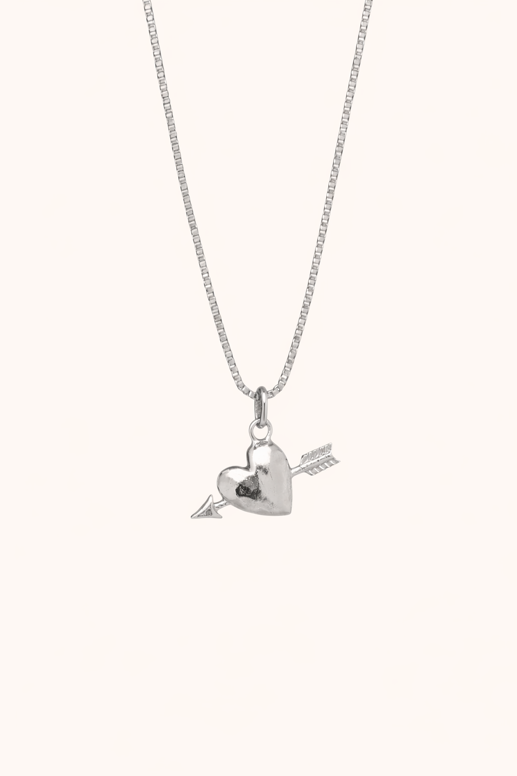 The Burning Hearts NECKLACE in silver, a puffed heart pierced by an arrow charm, hung from a box chain on a cream background.